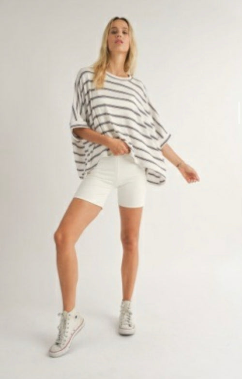Shows Sadie and Sage Model posing with Sailor Dolman Sleeve Knit Top, a white and navy striped t-shirt oversized style top. Model is also wearing white shorts and shoes. 
