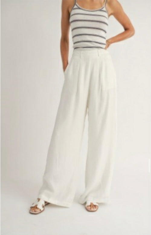 Model is wearing Sadie and Sage Heritage Wide Leg Pants, white linen pants with belt loops and pockets, relatively loose. Model is also wearing a striped tank and sandals.